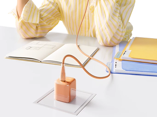 Person at a desk with notebooks, using a peach compact wall charger plugged into a desk outlet with a braided cable