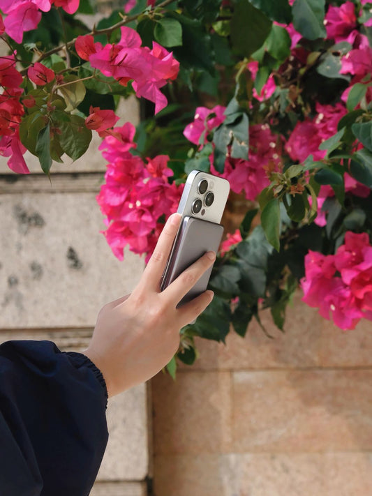 A hand holding a white smartphone with a silver magnetic power bank, posed against vibrant pink bougainvillea flowers.