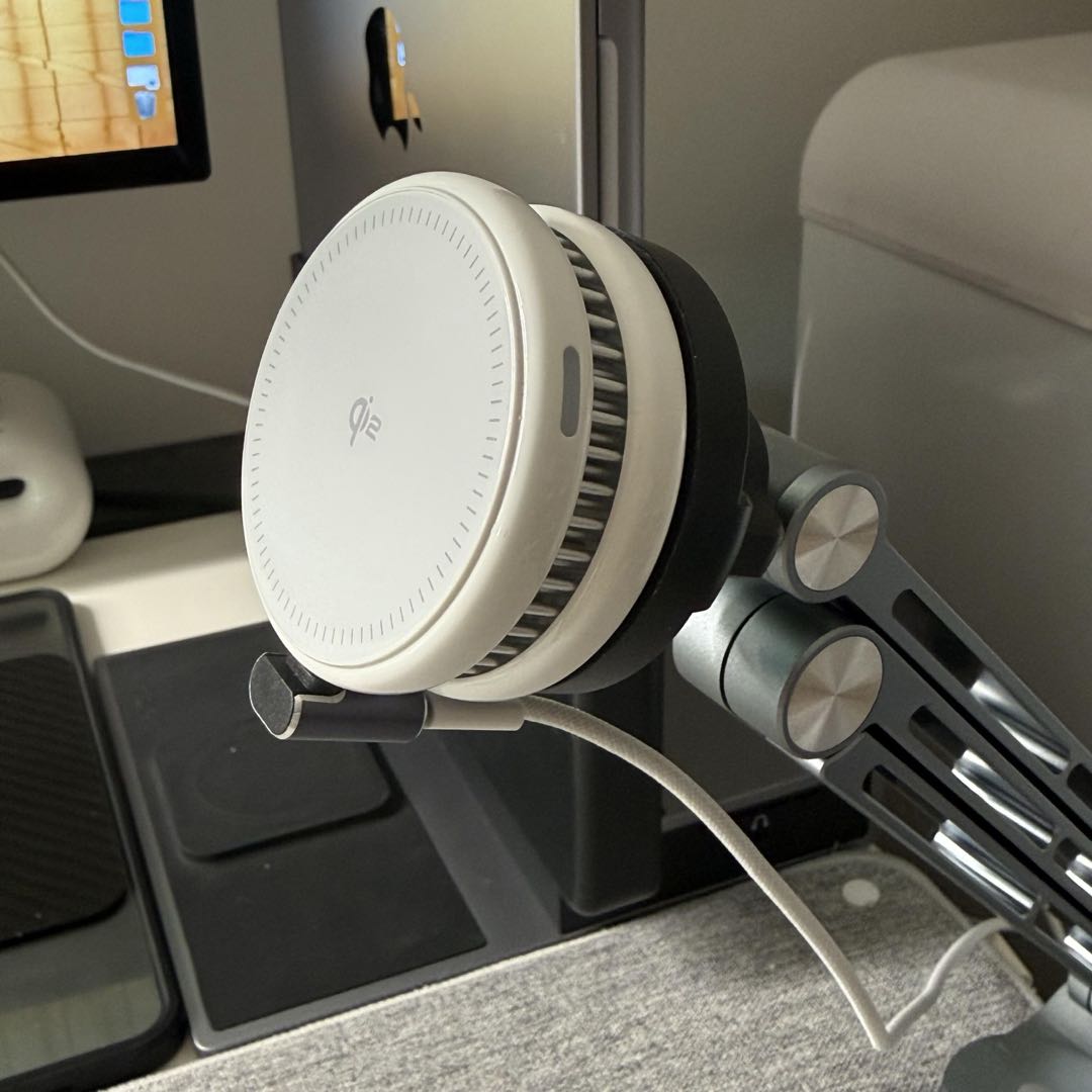 A white circular Qi2 wireless charger mounted on a gray adjustable metal arm at a desk setup.