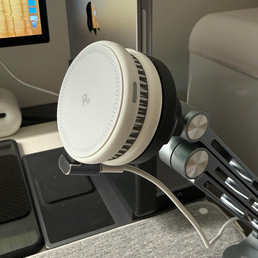 A white circular Qi2 wireless charger mounted on a gray adjustable metal arm at a desk setup.