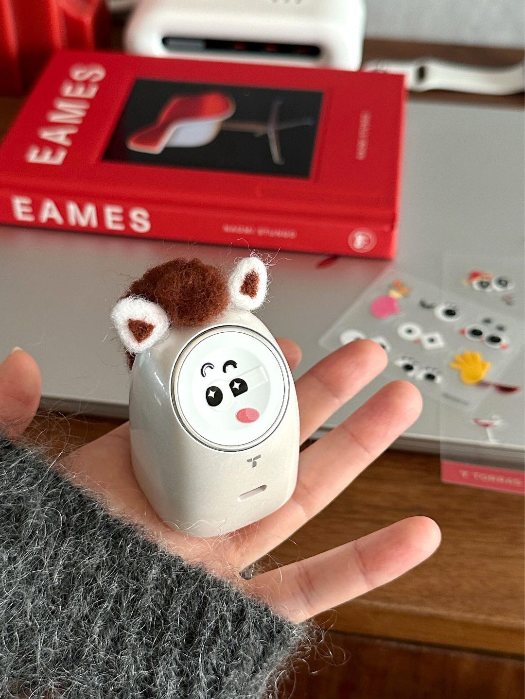 A hand holding a small, white electronic device decorated with fuzzy animal ears and a cartoon face sticker.