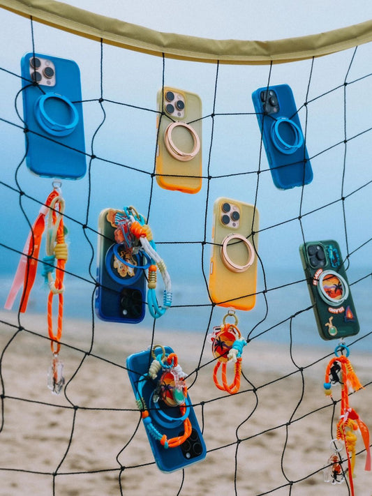 Colorful phone cases with ring holders and charms hanging on a beach net.
