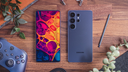 Smartphone comparison layout showing front and back of Samsung Galaxy phone on wooden desk with accessories and stylus nearby