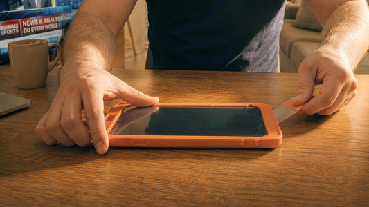Hands applying a screen protector to a smartphone secured in an orange alignment tray