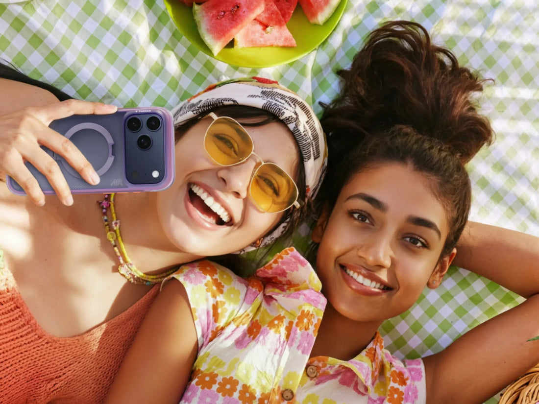 Overhead view of two smiling women lying on a checkered blanket, one holding a purple phone for a selfie.